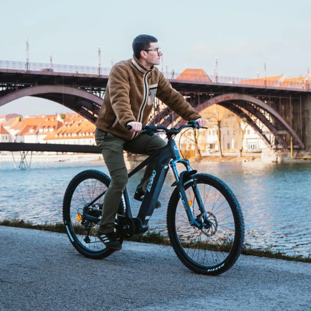 A person wearing a brown jacket is riding an electric bike Econo Gams SUV C6 along a riverbank with a bridge in the background