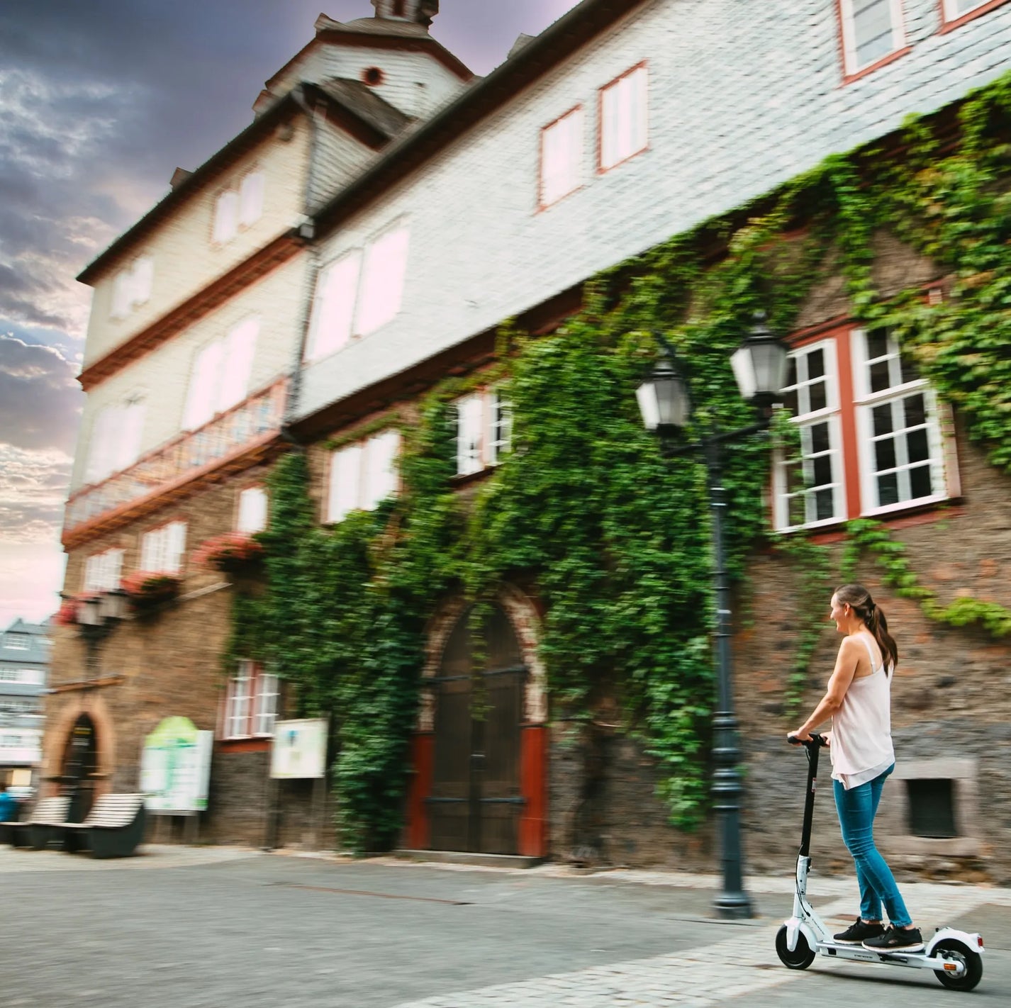 Električni romobil StreetBooster Vega – žena vozi bijeli romobil pored zgrade