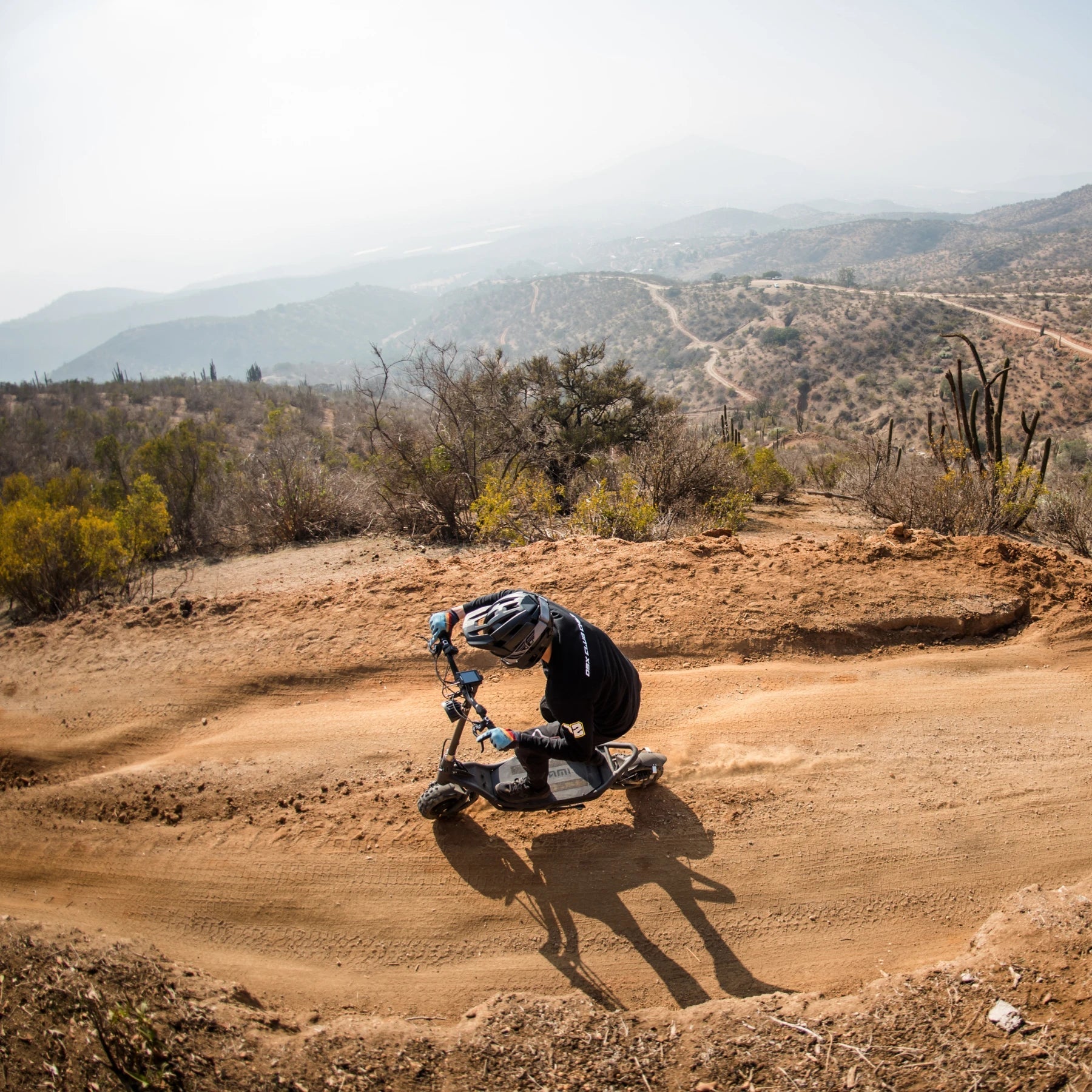Electric Scooter Nami Burn E 3 Max, rider maneuvering off-road on dirt trail, mountainous backdrop, shaded vegetation.